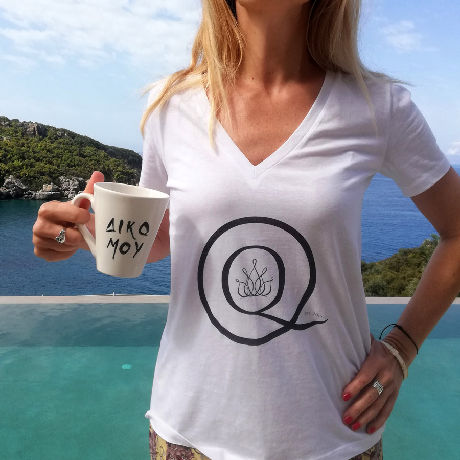 Woman wearing white t-shirt with black logo and holding white ceramic cup with black letters in front of a pool and the sea.